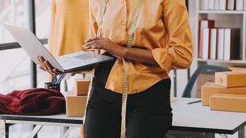 moda progettista l giovane asiatico donna Lavorando utilizzando computer portatile, tavoletta e sorridente mentre in piedi nel laboratorio rispondere su attività commerciale video