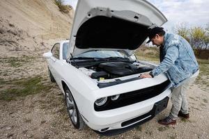 Handsome man in jeans jacket and cap is standing near his white muscle car with opening hood, check engine. photo