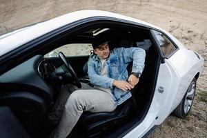 Handsome man in jeans jacket and cap sit and tie belt at his white muscle car. photo