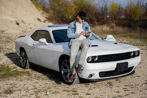 hermoso hombre en pantalones chaqueta y gorra sentar en capucha a su blanco músculo coche en carrera y mensajes de texto en teléfono. foto