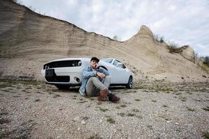 Handsome man in jeans jacket and cap is sitting near his white muscle car in career. photo