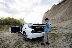 hermoso hombre en pantalones chaqueta y gorra abierto maletero desde su blanco músculo coche en carrera profesional. foto