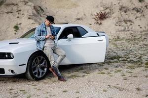 Handsome man in jeans jacket and cap is standing near his white muscle car in career and texting on phone. photo