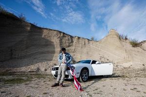 Handsome man in jeans jacket and cap with USA flag near his white american muscle car in career. photo