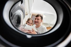 Man and girl view from washing machine inside. Father with daughter does laundry daily routine. photo