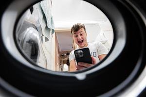 Man with mobile phone, view from washing machine inside. Male does laundry daily routine. photo