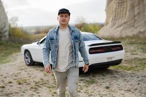 Handsome man in jeans jacket and cap walking near his white muscle car in career. photo