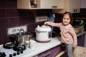 Baby girl prepare dinner at home kitchen with slow cooker and releases steam from the steamer. photo