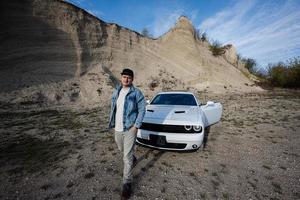 Handsome man in jeans jacket and cap is standing near his white muscle car in career. photo