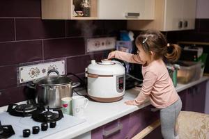 Baby girl prepare dinner at home kitchen with slow cooker and releases steam from the steamer. photo