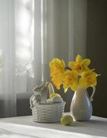 A nest with eggs in soft pastel colors and a vase of daffodils on a table by the window. Easter still life photo