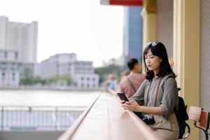 Young Asian woman backpack traveler using mobile phone in express boat pier on Chao Phraya River in Bangkok. photo