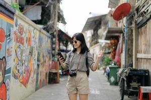 joven asiático mujer mochila viajero utilizando móvil teléfono, disfrutando calle cultural local Arte pared. viajero comprobación fuera lado calles foto