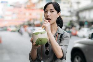 Happy young Asian woman backpack traveler drinking a coconut juice at China town street food market in Bangkok, Thailand. Traveler checking out side streets. photo