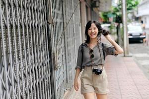 joven asiático mujer mochila viajero disfrutando calle cultural local sitio y sonrisa. viajero comprobación fuera lado calles foto