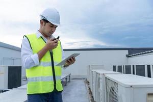 Asian maintenance engineer works on the roof of factory. contractor inspect compressor system and plans installation of air condition systems in construction. technology, online checking, mobile. photo