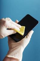 limpiando un smartphone con una servilleta amarilla estéril con guantes de goma sobre un fondo azul. foto