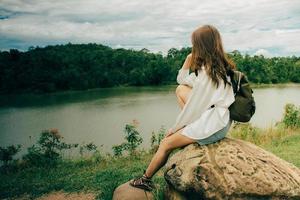 Young asian lonely woman while sitting at stone next to the river among mountains in summer. She was wearing a white shirt shoulder bag, thinking about her problems and felt very sad. photo