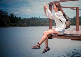 Beautiful Asian woman wearing glasses and backpack to travel the holidays. She was wearing a white shirt sitting on the bridge. using mobile phone to take pictures of river and mountains feel relaxed. photo