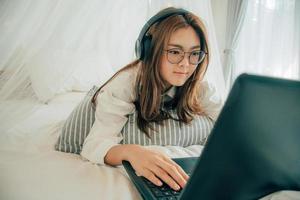 hermosa asiático joven mujer vistiendo lentes y auriculares escuchando música o acecho película con ordenador portátil en el blanco cama a hogar. bonito niña acostado abajo relajante y sonriente con cuaderno computadora. foto