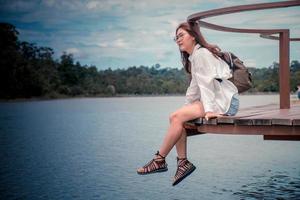 hermosa asiático mujer vistiendo lentes y mochila a viaje el vacaciones. ella estaba vistiendo un blanco camisa, sentado en el puente mirando a río montañas, el clima estaba excelente, sensación relajado. foto