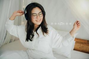 gracioso joven mujer con radiante sonrisa en gafas y vistiendo auriculares. escuchando a música canto y bailando en cama a hogar. ella usa blanco camisa. música amante ventilador pasatiempo concepto. música Nota icono. foto