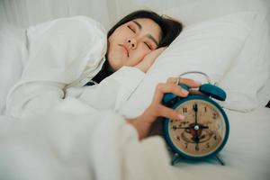 asiático hermosa mujer en blanco pijama torneado apagado alarma reloj mientras soñoliento en cama a su dormitorio de casa en vacaciones. niña es teniendo problema despertar arriba tarde en Mañana. insalubre dormir. selectivo atención foto