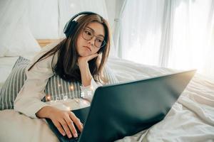 hermosa joven mujer vistiendo los anteojos y auriculares utilizando computadora portátil, buscando web, hojeada información, contacto, en línea compras y negocio concepto. teniendo lugar de trabajo a hogar. estilo de vida eso edad. foto