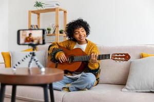 Blogger guitarist. African american girl blogger playing guitar talking to webcam recording vlog. Social media influencer woman streaming at home indoors. Music content creator broadcast tutorial. photo