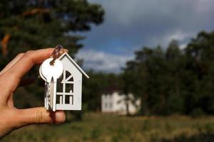 Wooden key chain and keys in hand on the background of an unfinished house. Dream of moving, the cottage is in a rural location and completion of the project. Space for text photo