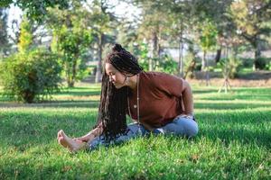 hermosa joven mujer con largo africano trenzas es haciendo yoga fuera de en un parque. concepto de sano estilo de vida. foto