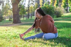 hermosa joven mujer con largo africano trenzas es haciendo yoga fuera de en un parque. concepto de sano estilo de vida. foto