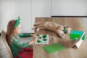 A little girl with a bandage on her head draws and cuts green shamrocks for St. Patrick's Day at a table at home in the kitchen, next to her is her beautiful cat with a green bow tie around his neck photo