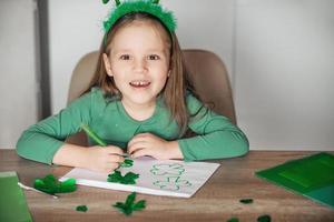 A little cheerful girl with a bandage on her head draws and cuts green shamrocks for St. Patrick's Day at her table at home. photo