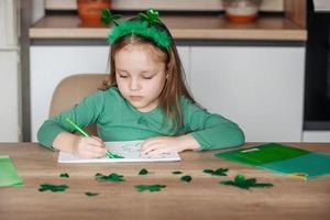 A little girl with a shamrock headband draws and cuts green shamrocks for St. Patrick's Day at her table at home photo