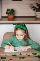 A little girl with a shamrock headband draws and cuts green shamrocks for St. Patrick's Day at her table at home photo