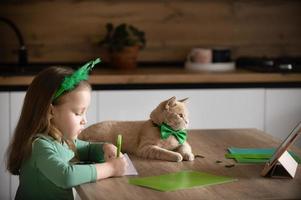 A little girl with a bandage on her head draws and cuts green shamrocks for St. Patrick's Day at a table at home in the kitchen, next to her is her beautiful cat with a green bow tie around his neck photo