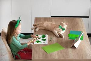 A little girl with a bandage on her head draws and cuts green shamrocks for St. Patrick's Day at a table at home in the kitchen, next to her is her beautiful cat with a green bow tie around his neck photo