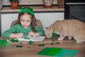 un pequeño niña con un vendaje en su cabeza sorteos y cortes verde tréboles para S t. patrick's día a un mesa a hogar en el cocina, siguiente a su es su hermosa gato con un verde arco Corbata alrededor su cuello foto