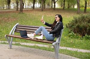 A young girl is sitting on a bench with a phone and laptop and taking a selfie photo