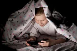 A little boy lies under a blanket at night playing on the phone photo