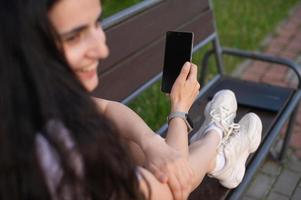 The girl is sitting with her feet on the bench, a watch on her hand, holding a phone in her hands photo