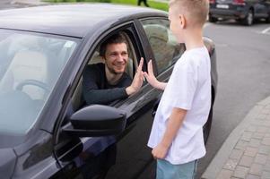 un linda niño acompaña un contento padre a trabajar. papá es sentado en el coche. dar cinco foto