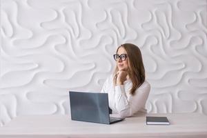 A young woman sits at her desk with a laptop and a diary and looks away photo