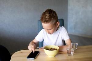 un linda chico es teniendo desayuno y mirando a el teléfono. un vaso de agua foto