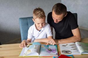 A cute boy is sitting at the table with his dad and watching a book about butterflies photo