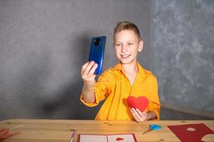 A cute boy is sitting at a table and making Valentine's Day cards. Taking selfies photo