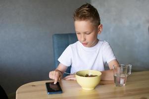 un linda chico es teniendo desayuno y mirando a el teléfono. un vaso de agua foto