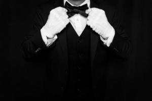 Portrait of Butler or Waiter in Black Vest and White Gloves Straightening Bow Tie on Black Background. Concept of Service Industry and Professional Hospitality photo