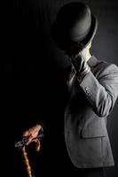 retrato de empresario en oscuro traje quitándose jugador de bolos sombrero en negro antecedentes. Clásico estilo y retro Moda de clásico Inglés Caballero. foto